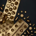 A striking overhead shot of golden-colored empty and partially filled blister packs, along with loose gold pills and capsules, scattered on a dark, almost black surface.