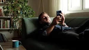 A man with glasses and a beard lies on a dark green sofa, looking at his smartphone. A blue mug is visible on a table nearby, and a plant and bookshelf are in the background.