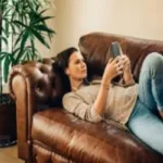 A woman lies on a brown leather sofa looking at her phone with headphones in, next to a green potted plant.