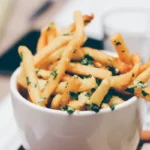 A white bowl overflowing with golden, crispy French fries sprinkled with green herbs.
