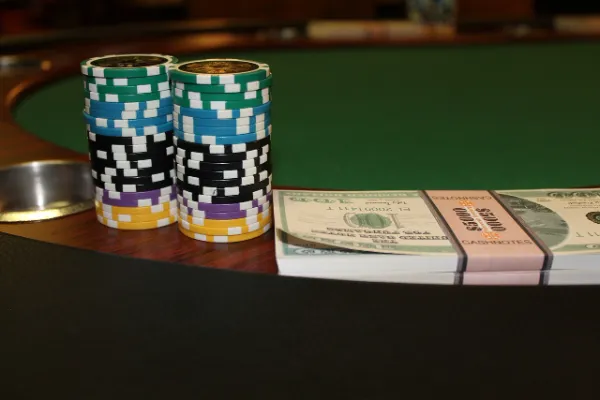 Two tall stacks of colorful casino chips (green, white, blue, black, purple, and yellow) next to a bundle of United States hundred-dollar bills wrapped with a pink "CASHNOTES $5000" band, all sitting on a wooden table with a green felt surface visible in the background.