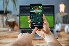 A person's hands holding a smartphone displaying a live sports betting app, with a laptop showing a sports game in the background, set on a wooden desk with a drink and headphones.