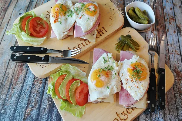 Two wooden boards, each with a slice of bread topped with ham, a fried egg, and herbs, accompanied by lettuce, tomato slices, and a small bowl of pickles.