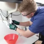 A plumber with safety glasses is shown working on pipes under a sink.