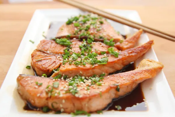 A white rectangular plate with several pieces of glazed salmon teriyaki, garnished with sesame seeds and chopped green onions, and chopsticks resting on the plate symbolizing online food trends jalbiteblog.