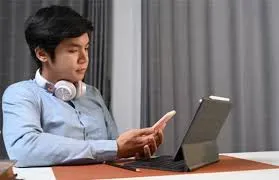 Person looking at a phone while working on a tablet with headphones around their neck.
