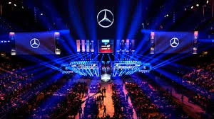 A vast esports arena illuminated with blue lights, showcasing a prominent Mercedes-Benz logo above the main stage and projected onto screens. A large audience fills the seating area, demonstrating the scale of the event and the presence of a major automotive sponsor.