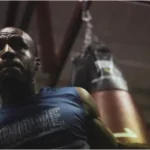 A boxer, possibly Evander Holyfield, wearing a blue training shirt and hand wraps, wipes his brow while standing near a hanging punching bag in a dimly lit gym.