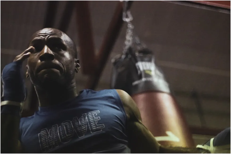 A male boxer with a serious expression, wearing a blue shirt and hand wraps, stands near a punching bag in a gym.