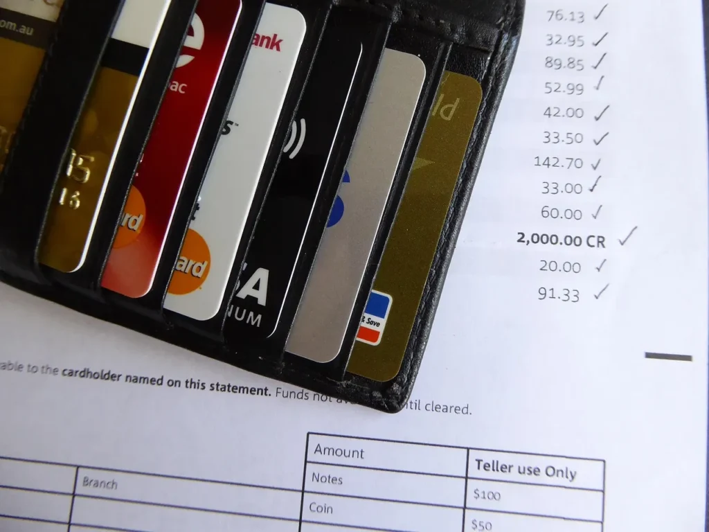 A close-up of various credit and debit cards partially visible in a black leather wallet, resting on top of a financial statement or bill with numbers and checkmarks visible.