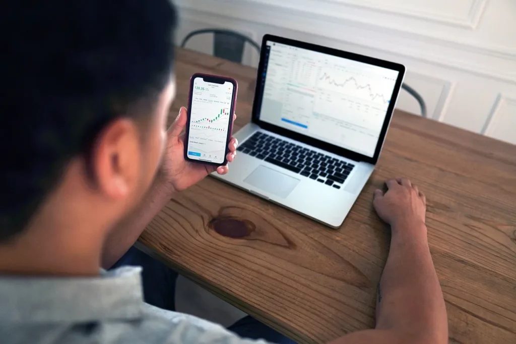 An individual using a smartphone to view stock market charts and financial data, with a laptop displaying similar information in the background, illustrating digital finance and investment analysis.