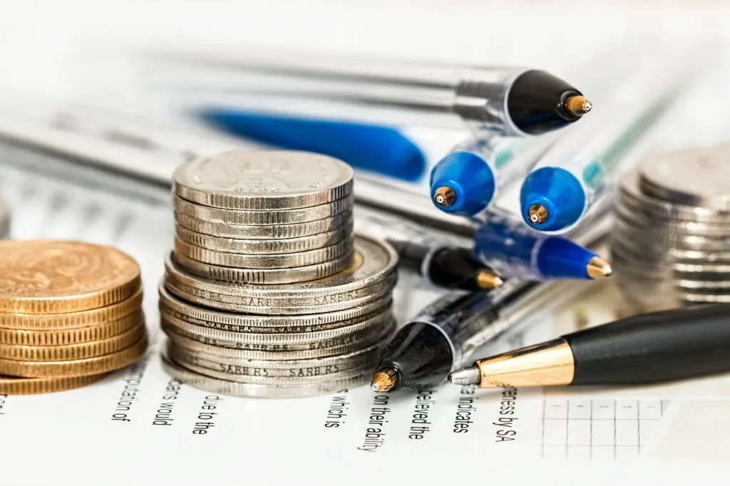 Stacks of coins alongside several ballpoint pens on a document with text.