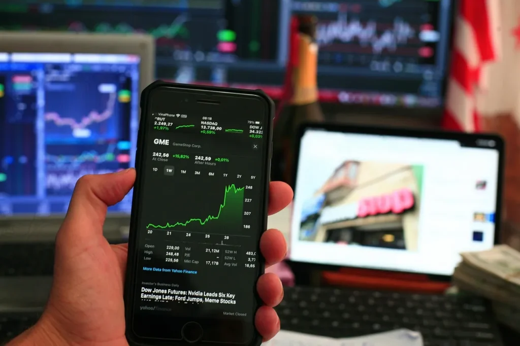 A hand holding a smartphone displaying a green upward-trending stock chart, with other computer screens showing financial data and a partial view of an American flag in the background, symbolizing market activity and tech-driven investment.
