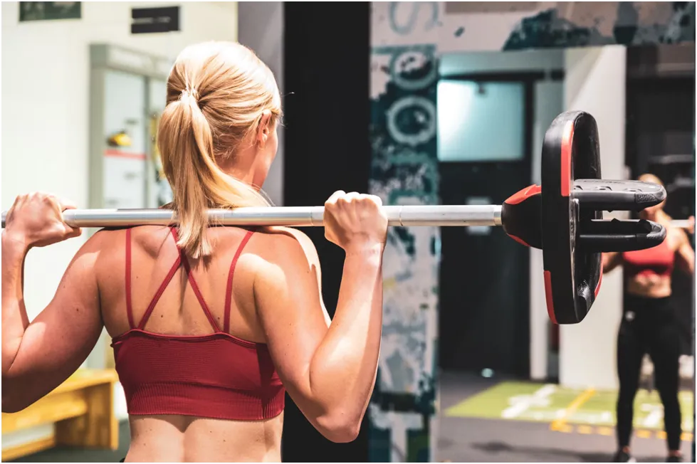 A woman with blonde hair tied in a ponytail holds a barbell across her shoulders, viewed from behind, in a gym setting.