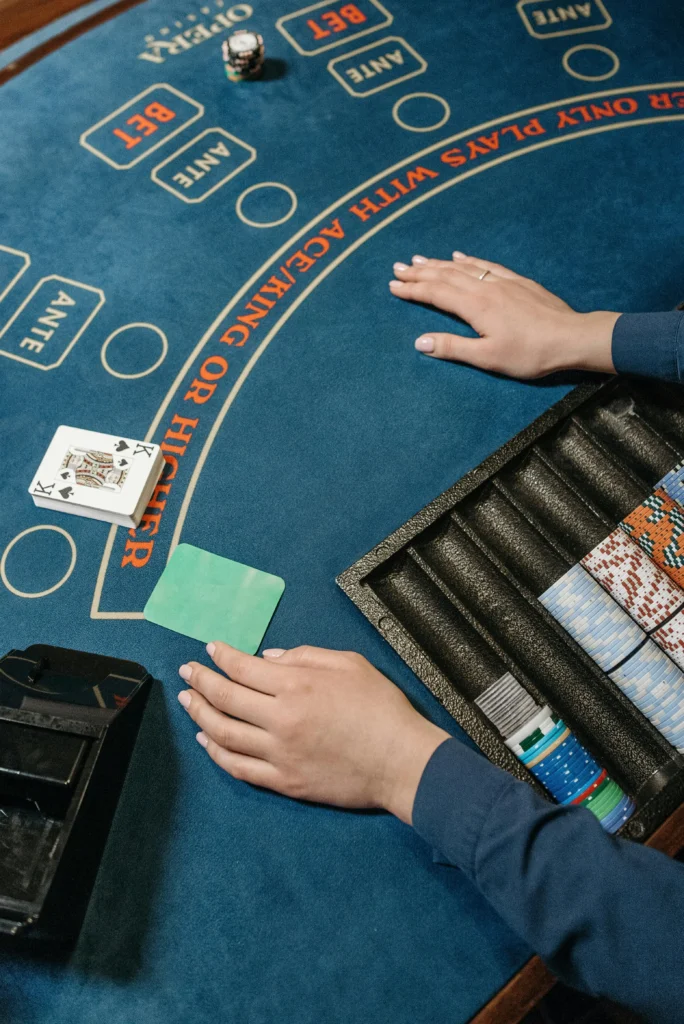 A top-down view of a blue Baccarat table, featuring a dealer's rack of chips, a hand resting on the felt, and a King card visible, indicating the start or middle of a casino game.