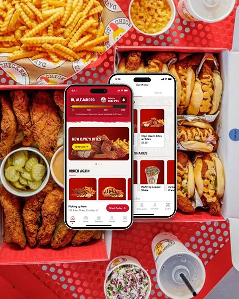 An overhead view of two open Dave's Hot Chicken catering boxes overflowing with fried chicken tenders, sliders, crinkle-cut fries, pickles, and mac and cheese, overlaid with two smartphones displaying the Dave's Hot Chicken mobile ordering app, demonstrating the ease of ordering from the Dave's Hot Chicken Menu.