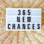 A white lightbox sign with black letters on a textured, light brown woven background. The sign reads "365 NEW CHANCES."