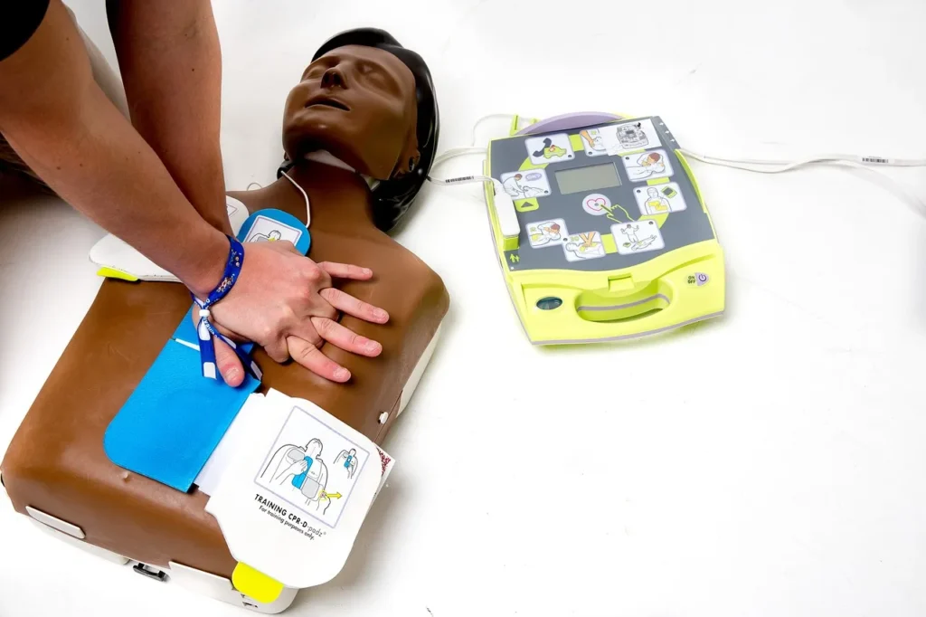 A person performing chest compressions on a CPR training mannequin with AED pads attached, next to a Zoll AED trainer device.