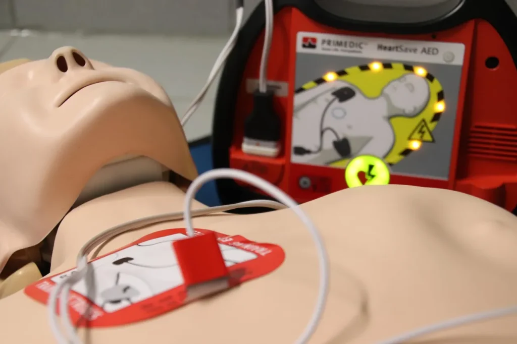 A close-up view of a CPR training mannequin's chest with an AED pad attached. Wires lead to a red AED trainer device in the background.