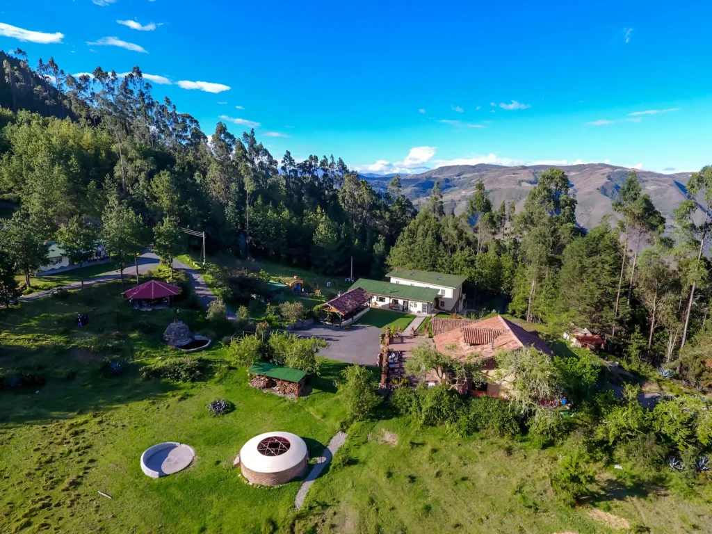 An aerial view of an Ayahuasca retreat center nestled in a lush green valley with mountains and tall trees under a bright blue sky in Ecuador.
