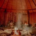 A group of people sitting in a circle around a central fire pit inside a traditional, round hut or maloka.