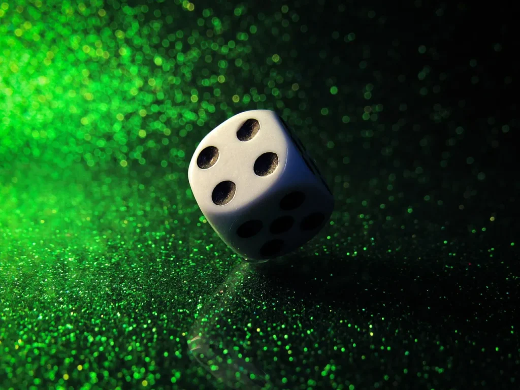 A single white casino die with black pips, resting on a sparkling green surface with a dark gradient, illuminated by a spotlight effect.