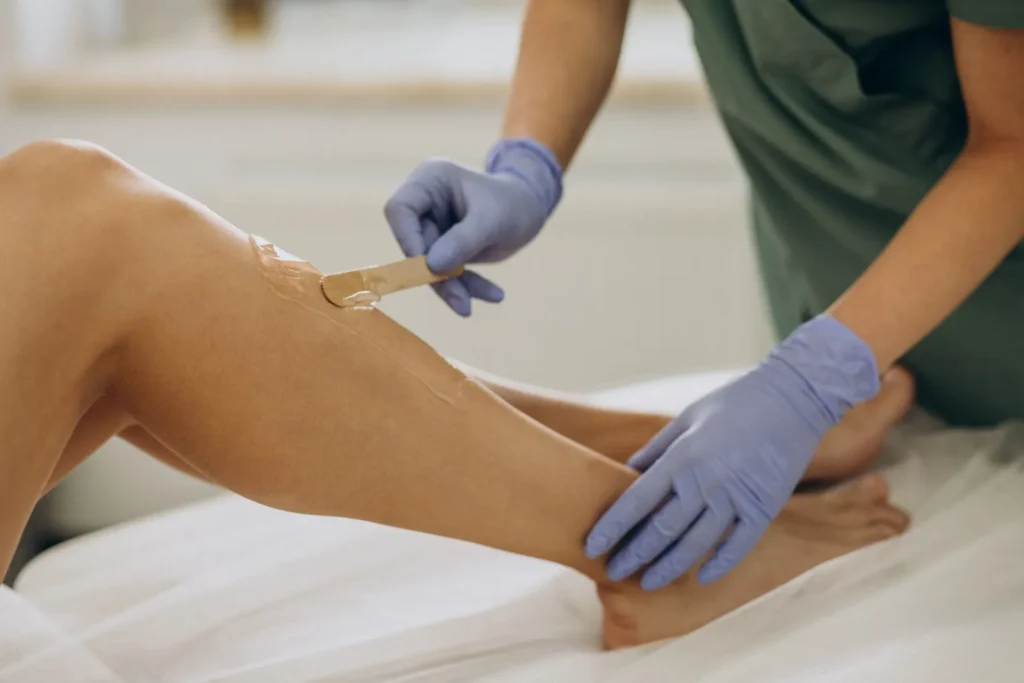 A close-up of an esthetician in blue gloves applying wax to a client's leg with a wooden stick.