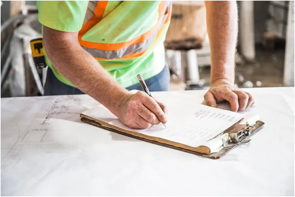 A general contractor in a safety vest reviewing blueprints on a clipboard.