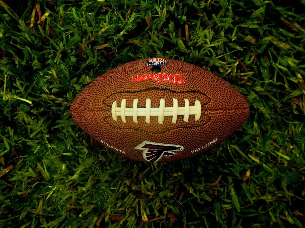 A close-up, overhead view of a brown leather American football with the Atlanta Falcons logo lying on thick, dark green grass, symbolizing the start of the NFL season.