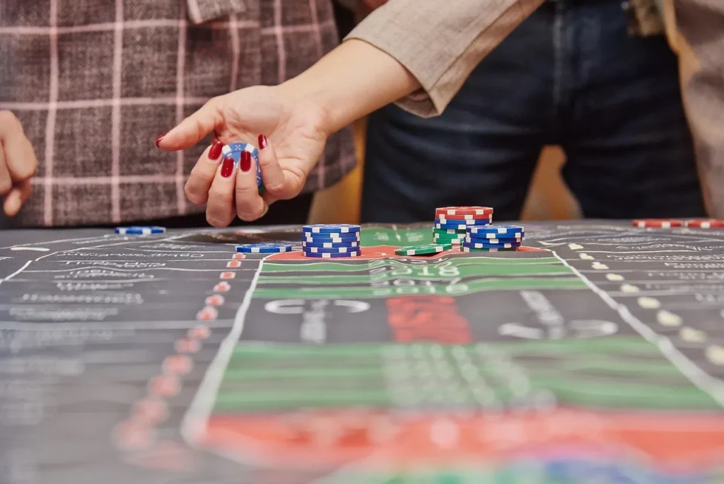 A person's hand with red-painted fingernails placing blue and white casino chips onto a marked casino table, with stacks of other chips visible, representing the act of betting and engagement in casino games.