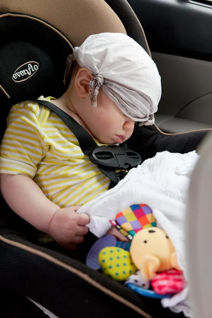 A baby in a car seat with a cloth covering their eyes, buckled in with a car seat harness.
