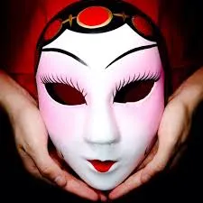 A close-up shot of a pair of hands holding a refined Chinese opera mask. The mask has a predominantly white face with soft pink accents on the cheeks, and thin, graceful black eyebrows and eyelashes. The lips are painted a delicate red, and a decorative red and black headdress with gold trim sits atop the mask. The mask symbolizes a gentle or scholarly female character.