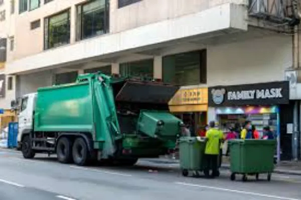 A large green garbage truck is parked on a city street, actively collecting waste from two green commercial dumpsters, with a worker in a yellow vest assisting.