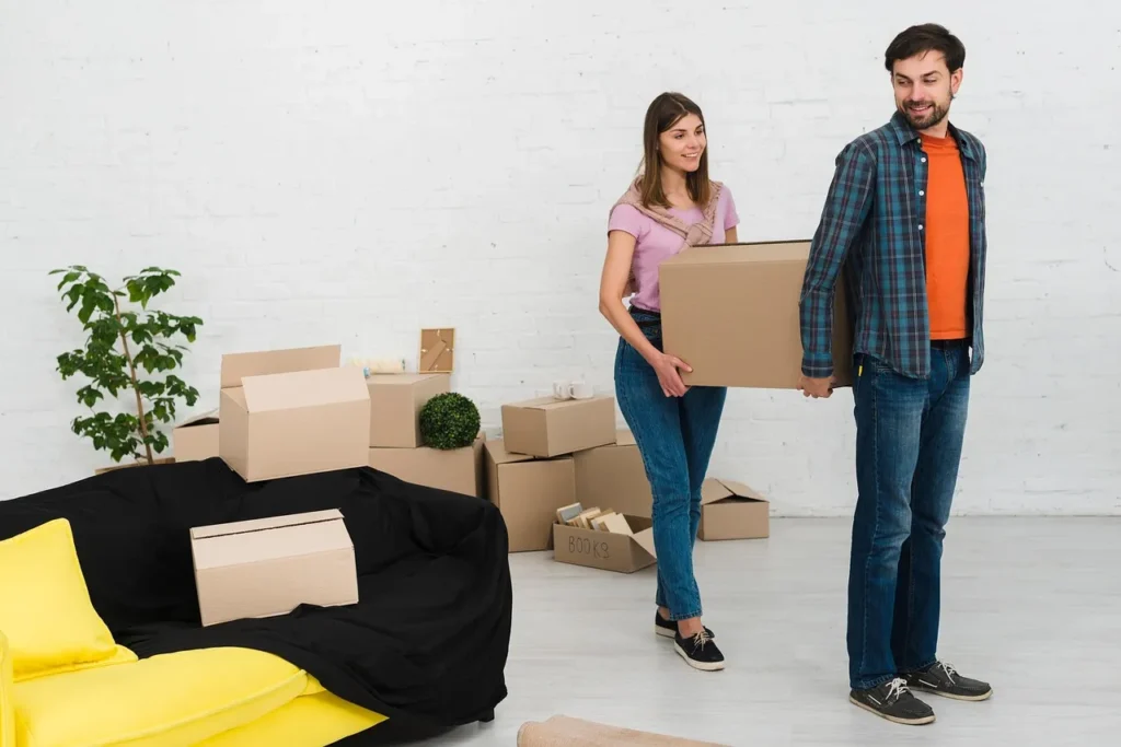A smiling couple carrying a large cardboard box into a white-walled room with a black couch and several unpacked moving boxes piled up in the background.