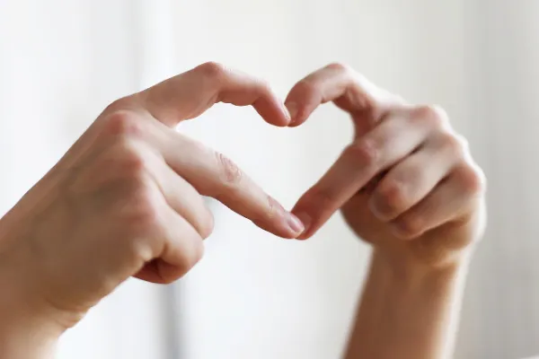 Two hands form a heart shape with their fingers, symbolizing love, care, and compassion in a relationship, set against a soft, light background.