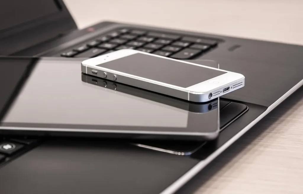 A silver smartphone is resting on a black tablet, which in turn is placed on top of a dark laptop with a visible keyboard and trackpad, showcasing multiple digital devices.