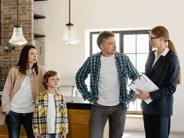 A real estate agent in a suit holds a clipboard, speaking with a family (a couple and their child) in what appears to be a new home or property.