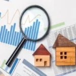 A magnifying glass rests over financial charts and spreadsheets, with two small wooden house models beside it, symbolizing property market analysis.