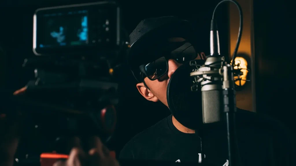 A person wearing a cap and sunglasses speaks into a large studio microphone, with a video camera visible in the background, symbolizing content creation, explanation, or broadcasting a viral trend.