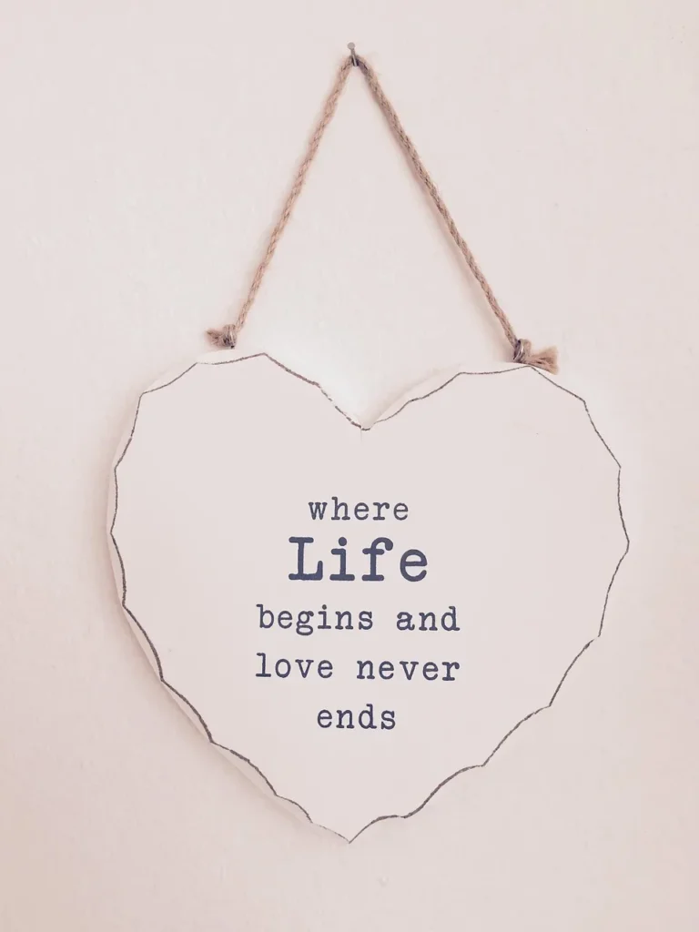 A white wooden sign, shaped like a heart and hanging from a twine string against a white wall, inscribed with the quote: "where Life begins and love never ends."