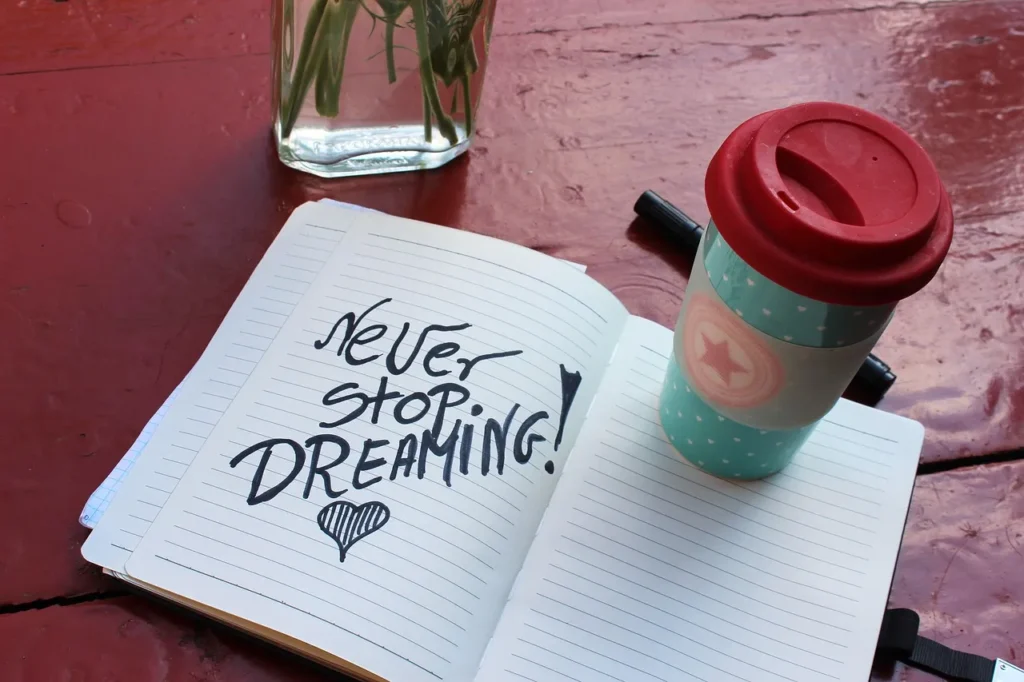 An open lined notebook resting on a red surface with the handwritten quote "Never stop DREAMING!" and a heart drawn underneath, next to a travel mug.