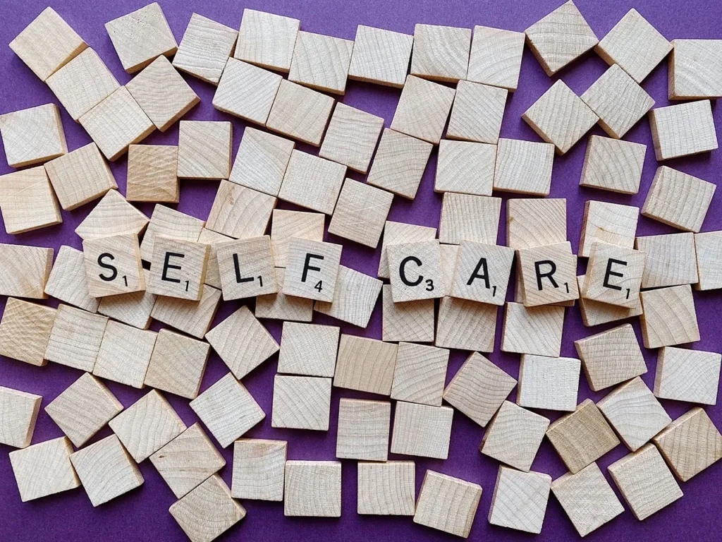 An overhead shot of numerous wooden Scrabble tiles scattered on a purple surface, with seven tiles spelling out "SELF-CARE."