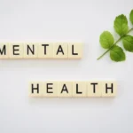 The words "MENTAL HEALTH" spelled out in two rows using ivory square tiles, positioned next to a fresh green leaf on a clean white background, symbolizing natural well-being.