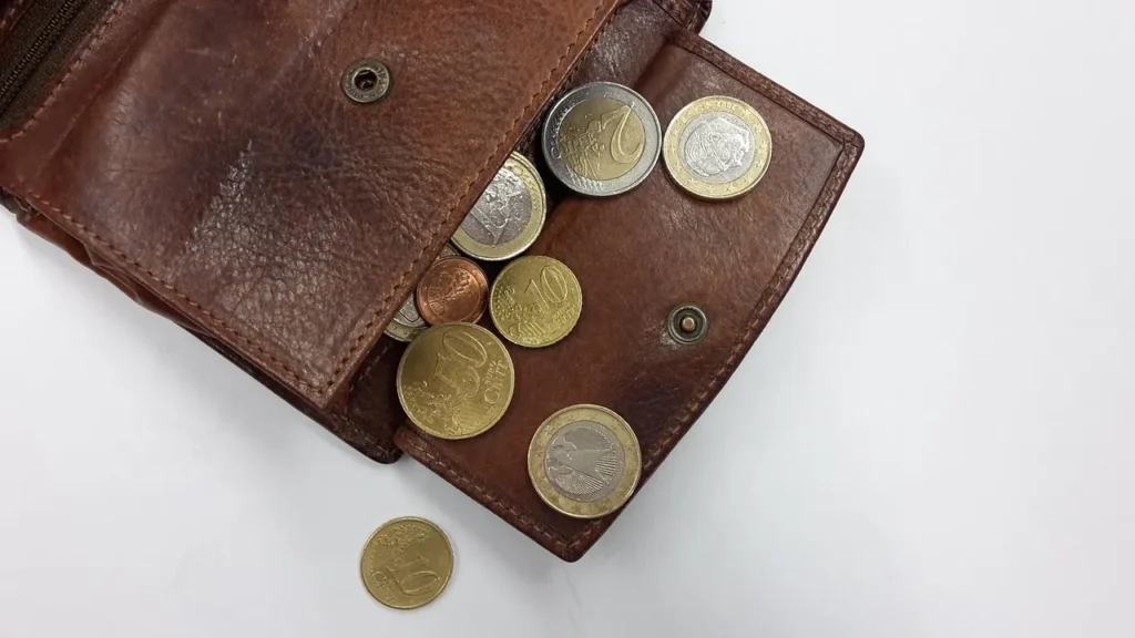 A close-up, overhead view of a brown leather wallet opened to show a collection of Euro coins (10 cent, 1 Euro, 2 Euro) spilled onto a white surface, symbolizing saving money and budgeting during a no-spend challenge.