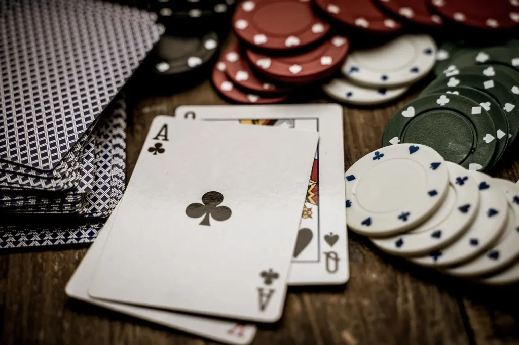 A close-up shot of two face-up cards (Ace of Clubs and Queen of Hearts) with stacks of red, green, and white poker chips and a deck of cards scattered on a dark wooden table.