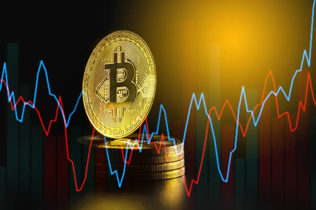 A large gold physical Bitcoin coin stands on a stack of other coins, centered in front of a volatile red and blue cryptocurrency trading graph on a dark background.