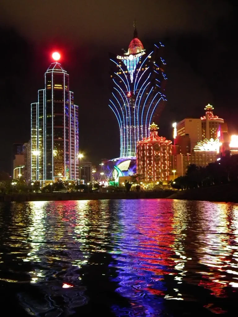 A dramatic, long-exposure photograph of the brightly lit, modern casino buildings of the Macau skyline at night, reflected in the dark water in the foreground.
