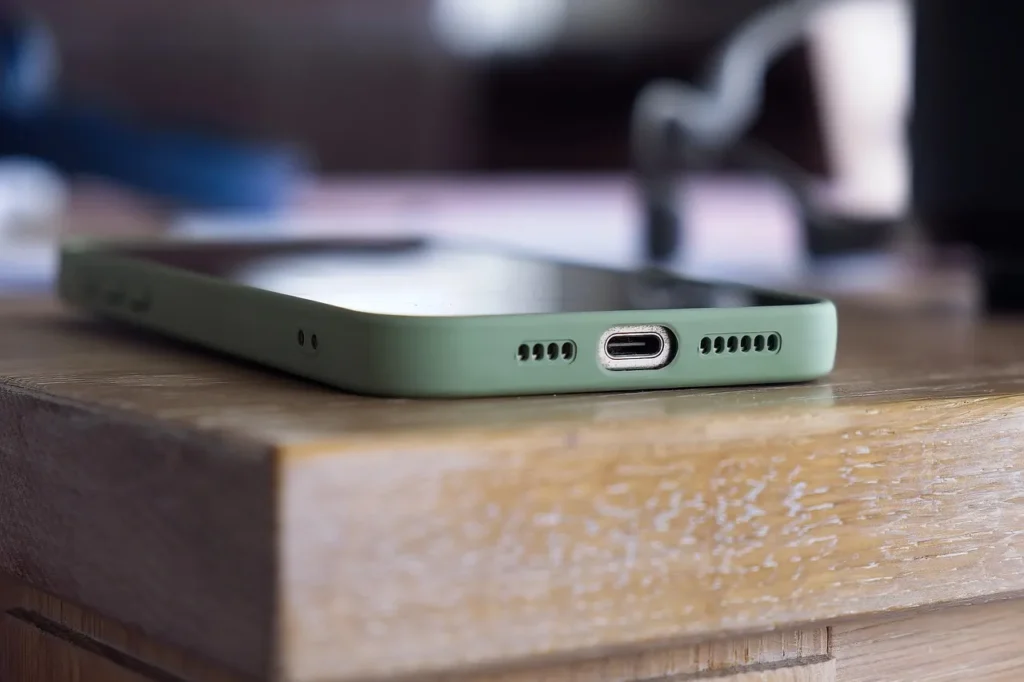 An extreme close-up of the bottom edge of a smartphone in a green case, showing the USB-C or Lightning port flanked by tiny speaker and microphone grilles, resting on a wooden surface.