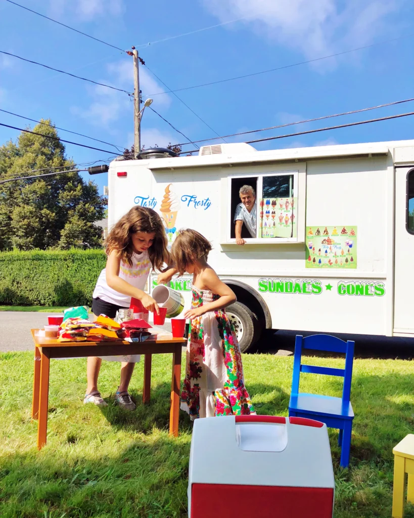 Two young girls in summer dresses standing at a small wooden table outside a white ice cream truck called "Tasty Frosty." One girl pours a drink into red cups while the ice cream man smiles from the truck window.
