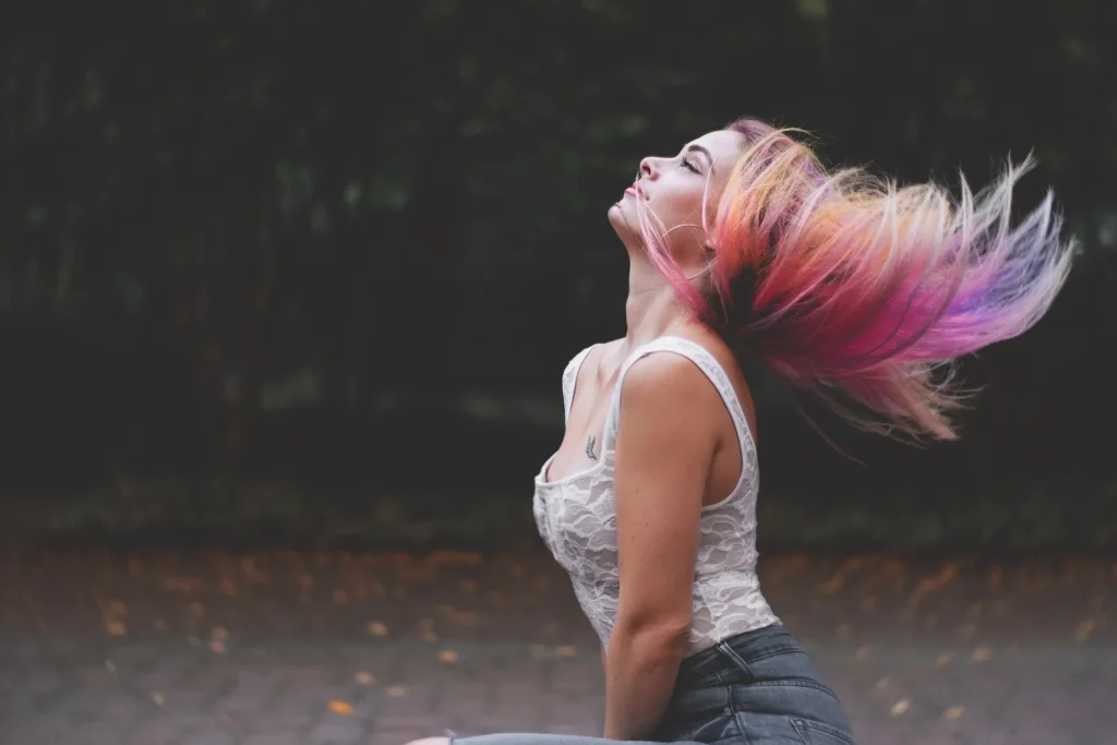 A woman with vibrant pink and orange hair tossing her head back in a motion blur, wearing a white lace bodysuit and grey jeans against a dark, out-of-focus park background.